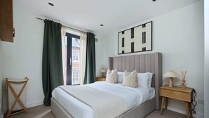A bedroom with a large bed, a bedside table with a lamp, a wall-mounted artwork, and a view through a window with curtains. - The Shoreditch Retreat - Inviting 2bdr Apartment (London)
