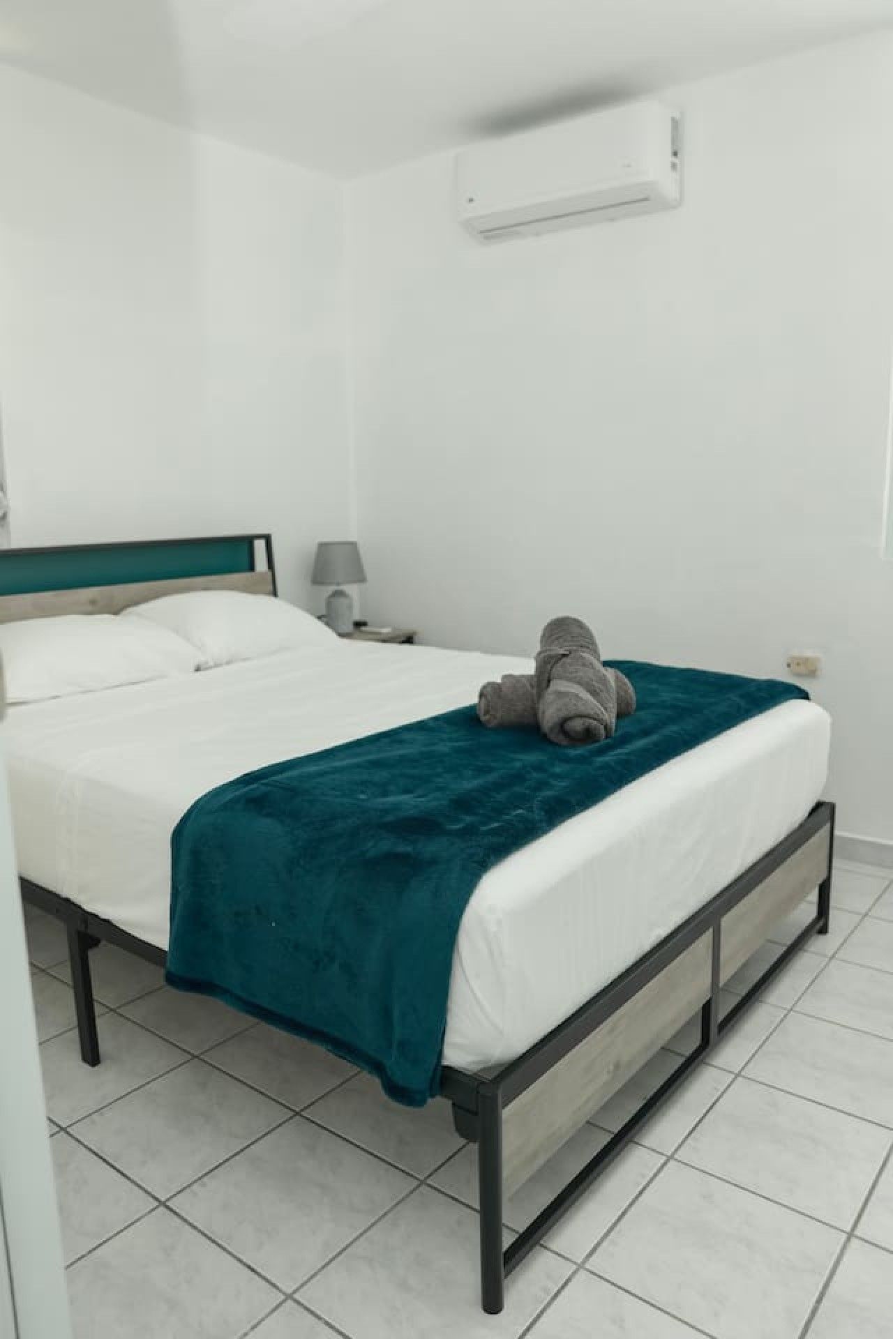 A single-bed room with a teal bedspread, white bedding, a grey towel, and a bedside lamp.