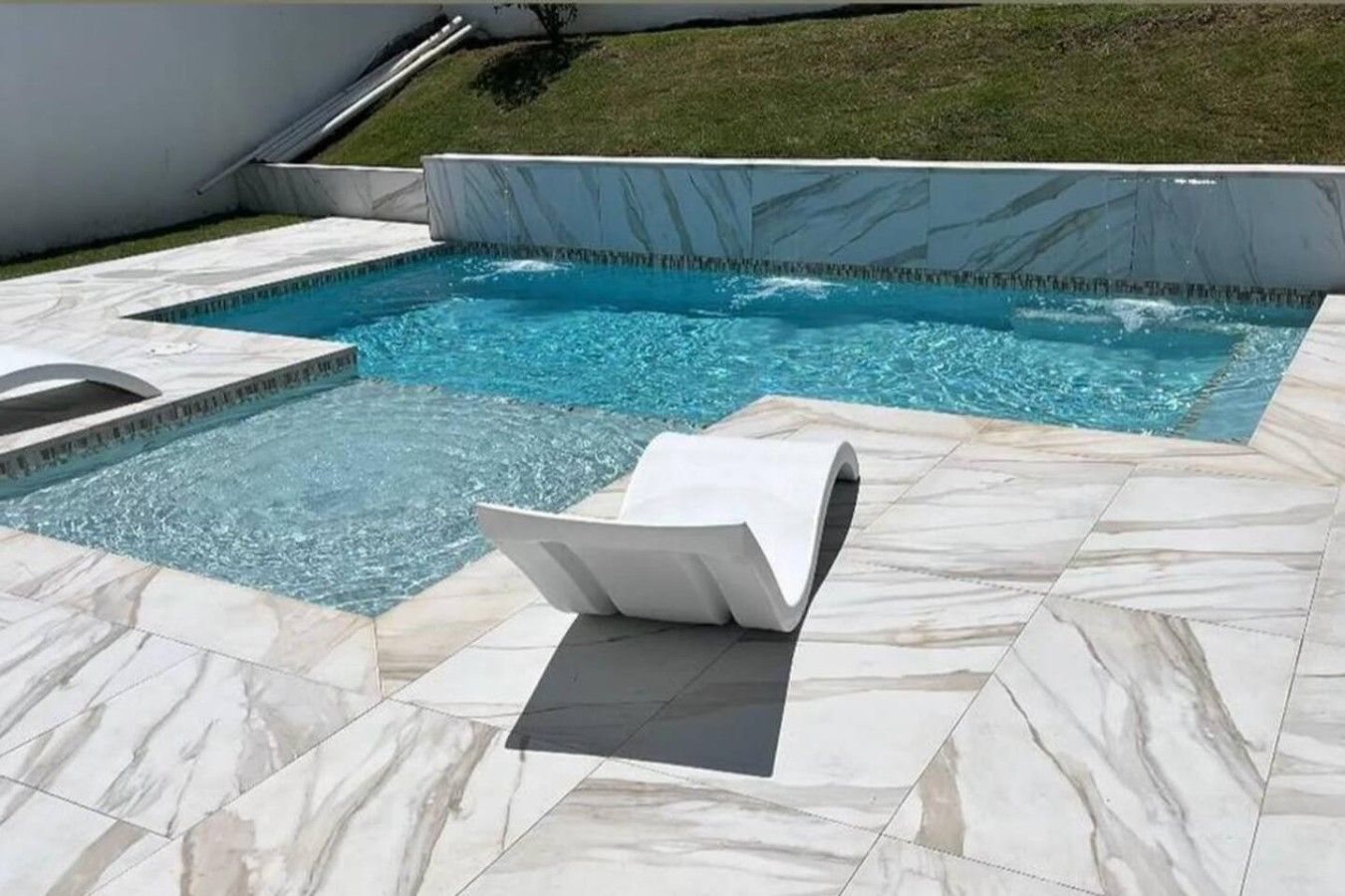 A swimming pool with a tiled deck, a lounge chair, and a small water feature.