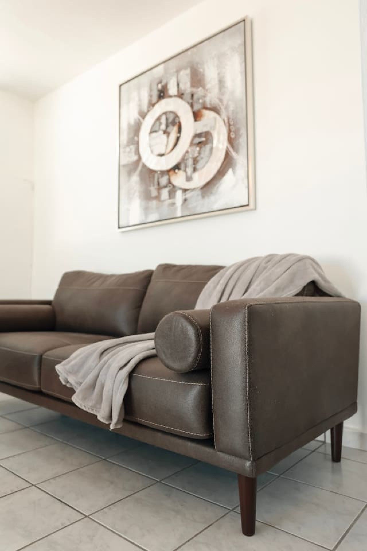 Un divano in pelle marrone con una coperta, posto in una stanza con pavimento piastrellato e un grande quadro astratto appeso al muro.