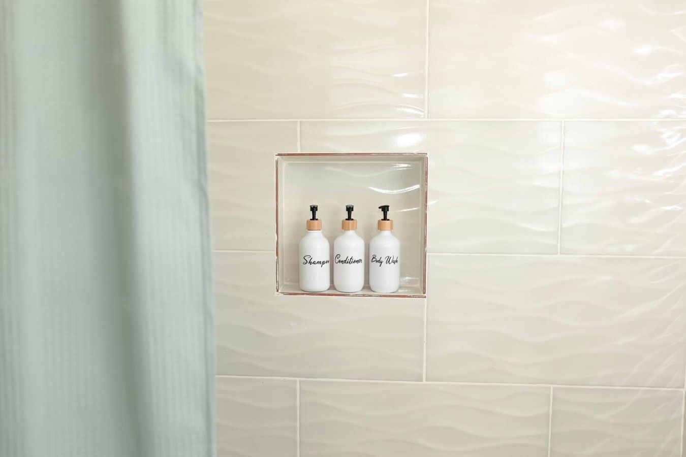 Een badkamer met drie flessen shampoo, conditioner en douchegel in een transparante glazen houder aan de muur.