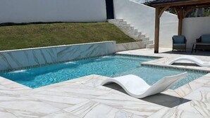 Ein Schwimmbecken mit einer Marmor-Terrasse, einer sanften Grasfläche und einem überdachten Sitzbereich mit Stühlen.