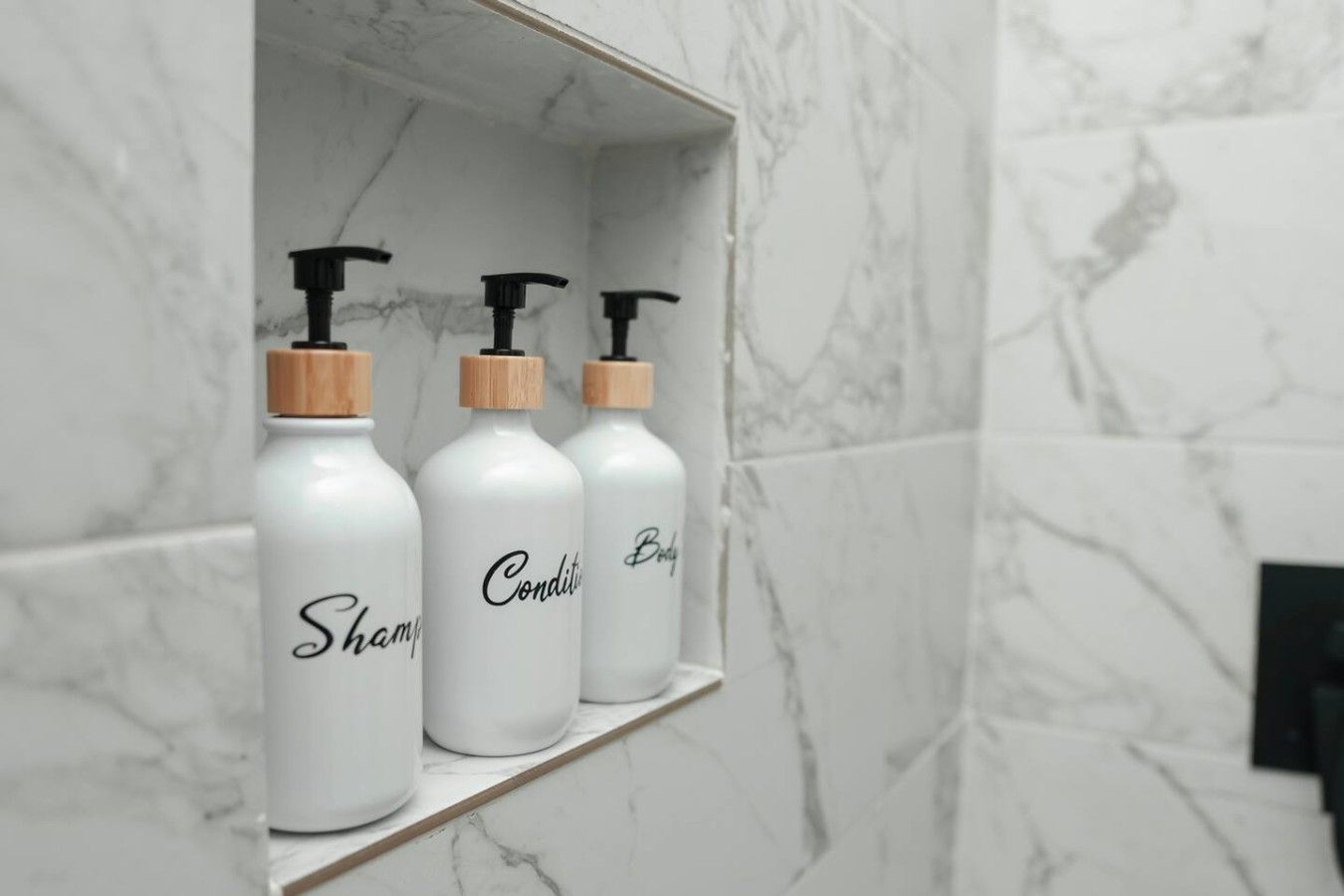 A bathroom shelf with three white bottles labeled 'Shampoo,' 'Conditioner,' and 'Body Wash.'