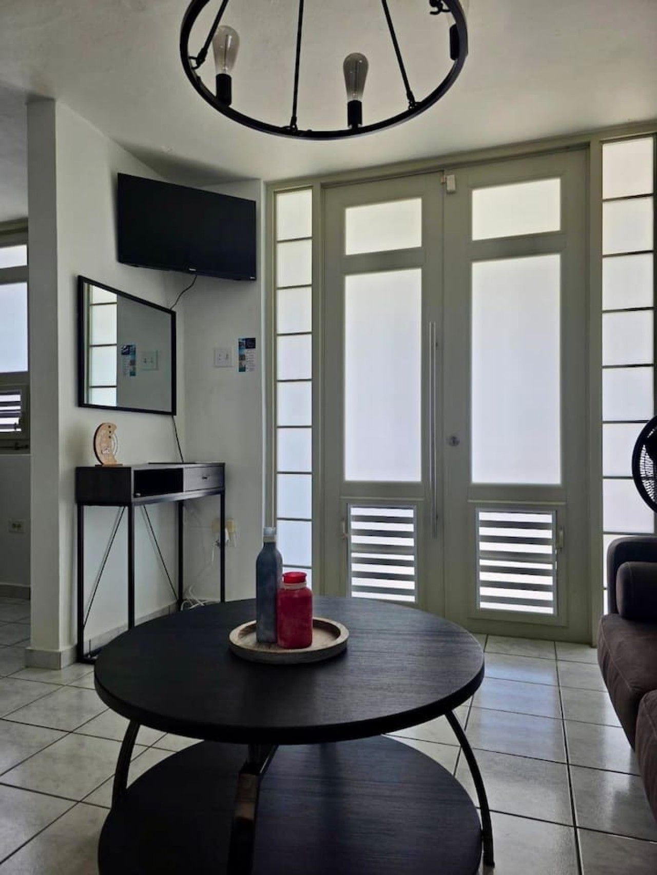A room with a round table, a flat-screen TV mounted on the wall, and a large window with blinds.