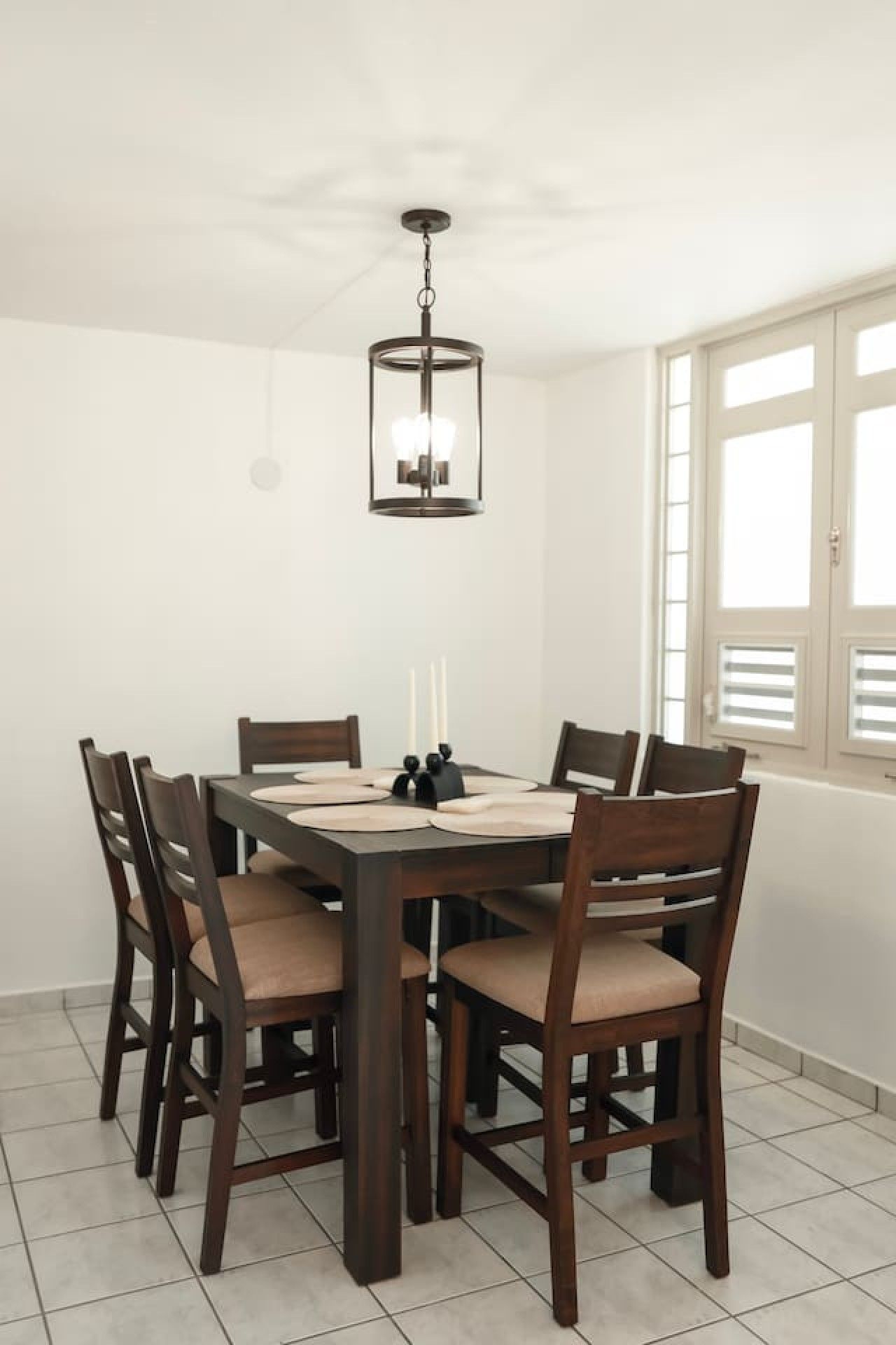 Un coin repas comprenant une table rectangulaire, six chaises, un luminaire suspendu et une fenêtre avec des stores.