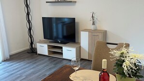 Ein Esstisch mit Platzsetzern, Weingläsern und einer Weinflasche. Im Hintergrund befinden sich ein Fernseher und Schränke.