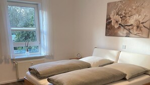 Ein Schlafzimmer mit einem Holzbett, weißer Bettwäsche, einem Fenster mit Vorhängen, einem Heizkörper und einem Bild an der Wand.