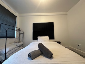 A neatly made bed with white linens and two rolled towels, a black headboard, a window with blinds, and a radiator.