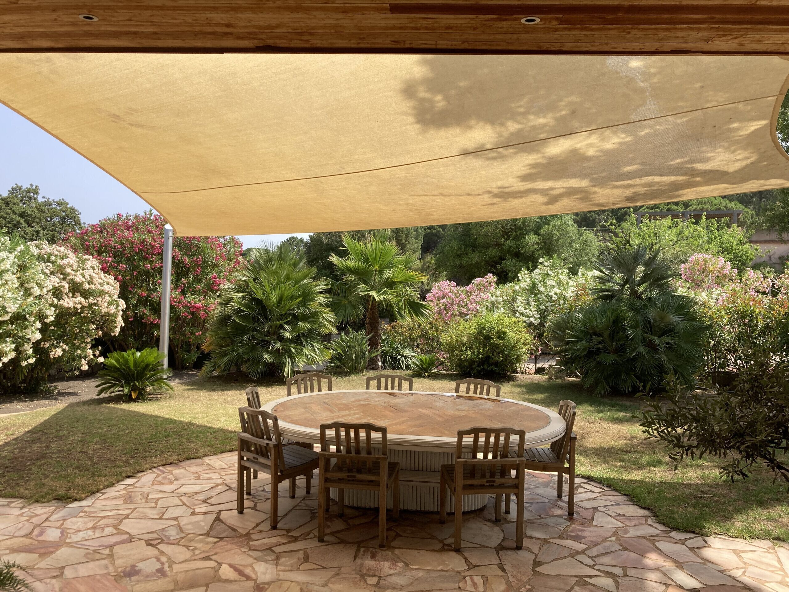 A stone patio with a round table and chairs, surrounded by lush greenery and flowering plants.