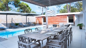 Un espace repas aménagé au bord de la piscine, avec une longue table en bois et des chaises, entouré de parasols et de fauteuils de relaxation.