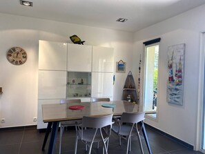 Un espace repas moderne avec une table à plateau en verre, deux chaises grises, un meuble blanc avec des portes en verre, une horloge murale et un poisson décoratif.