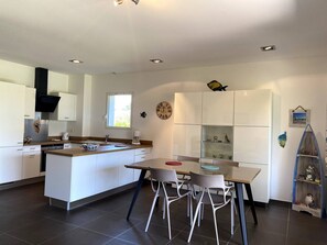 A modern kitchen with white cabinetry, a central island, and a dining area with chairs. - T3 face à la Mer by Pinarello Villa Services (Sainte-Lucie de Porto-Vecchio)