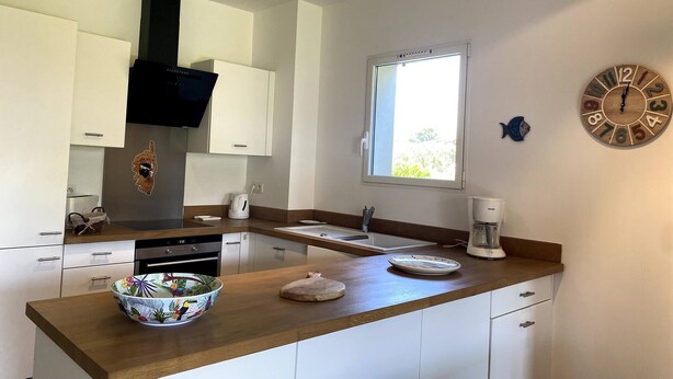 A modern kitchen with white cabinets, a black range hood, a window, and a wooden clock on the wall. - T3 face à la Mer by Pinarello Villa Services (Sainte-Lucie de Porto-Vecchio)