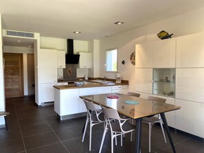 Une cuisine moderne avec des meubles blancs, un îlot central et un coin repas comprenant une table et des chaises en verre.