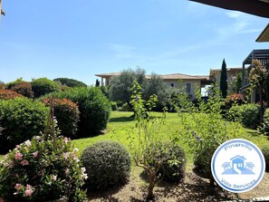 A villa with a well-maintained garden, a clear blue sky, and a logo for Pinarello Villa Services.