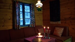 Interior acolhedor de cabana com parede de madeira, janela com cortinas estampadas, luminária pendente, mesa com vela acesa e sofá com almofadas.