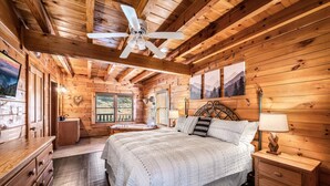A wooden cabin bedroom with a bed, nightstands, a ceiling fan, and a window with a view of the outdoors.