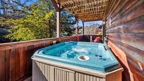 A hot tub under a wooden pergola with a wooden deck and a view of trees.