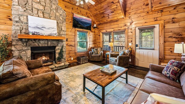 A cozy log cabin interior with a stone fireplace, leather furniture, and a ceiling fan.