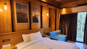 A hotel room with a large bed, a nightstand, a blue chair, and a view of the outdoors through a window. - TUPTUP - Resto & Resort (Cox's Bazar)