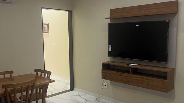 A living room with a TV mounted on the wall, a dining table with chairs, and a door leading to another room.