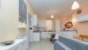 A modern kitchen with white cabinets, a dining area with a grey sofa, and a wall with a clock and decorative items. - Luvi Maison - Affitti Brevi Italia (Terrazzano)