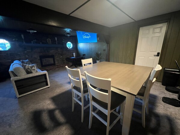 Un coin repas avec une table en bois et des chaises blanches, une cheminée en pierre et un téléviseur fixé au mur.