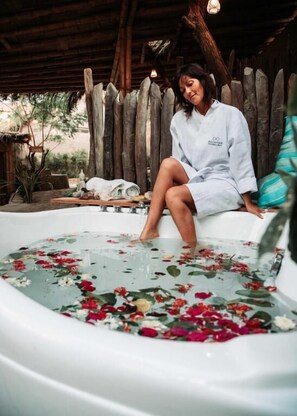 A person sitting in a hot tub with floral petals floating on the water, surrounded by wooden structures and greenery. - AguaMar Vichayito (Los Organos)