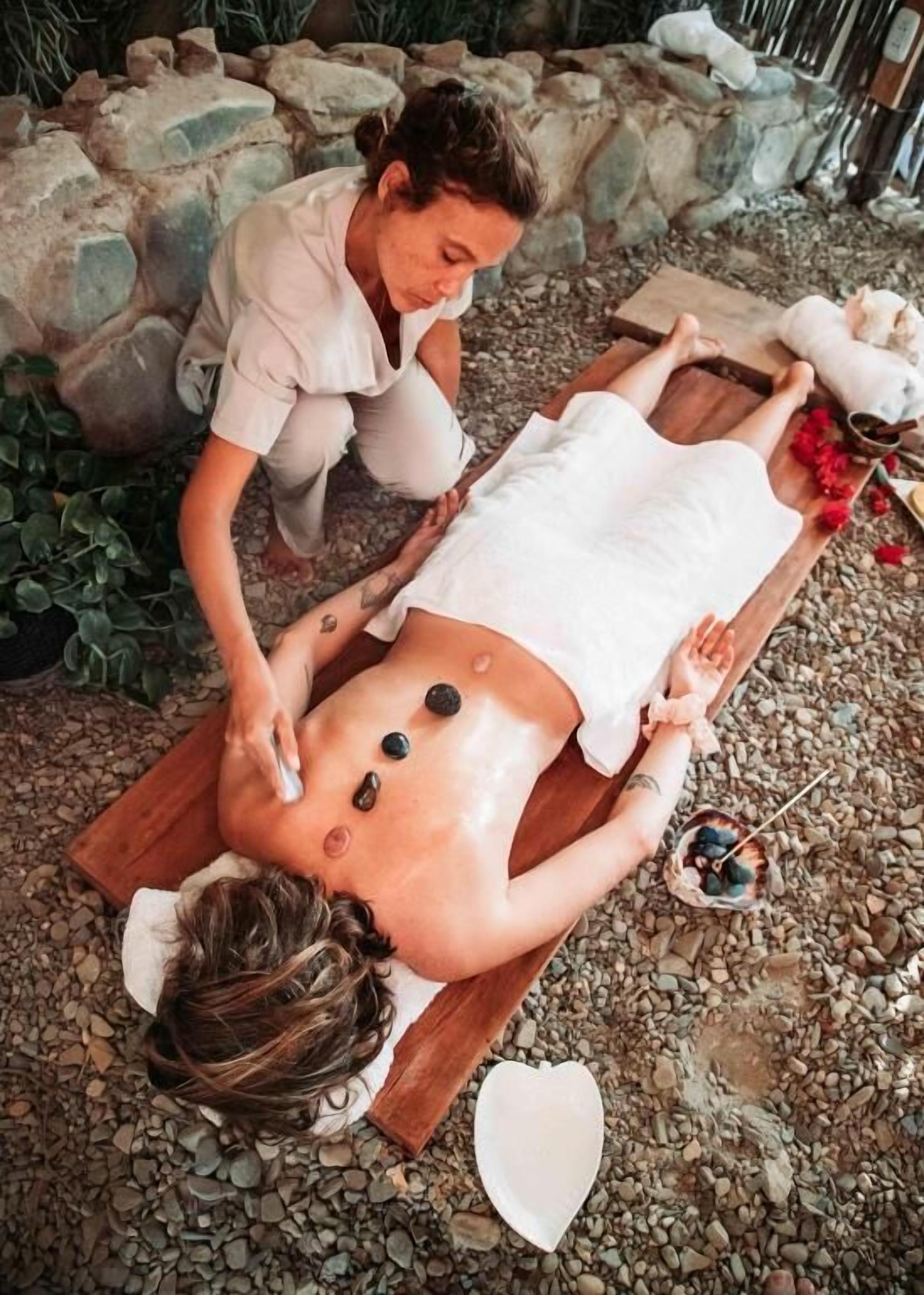 Una persona recibiendo un masaje con piedras al aire libre, atendida por un terapeuta.