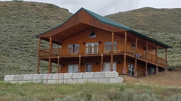 A two-story wooden cabin with a balcony, set against a mountainous backdrop.