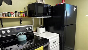 Cucina con piano cottura nero, forno bianco e frigorifero nero. Sopra il piano cottura c'è un forno a microonde, e una mensola con diverse spezie.