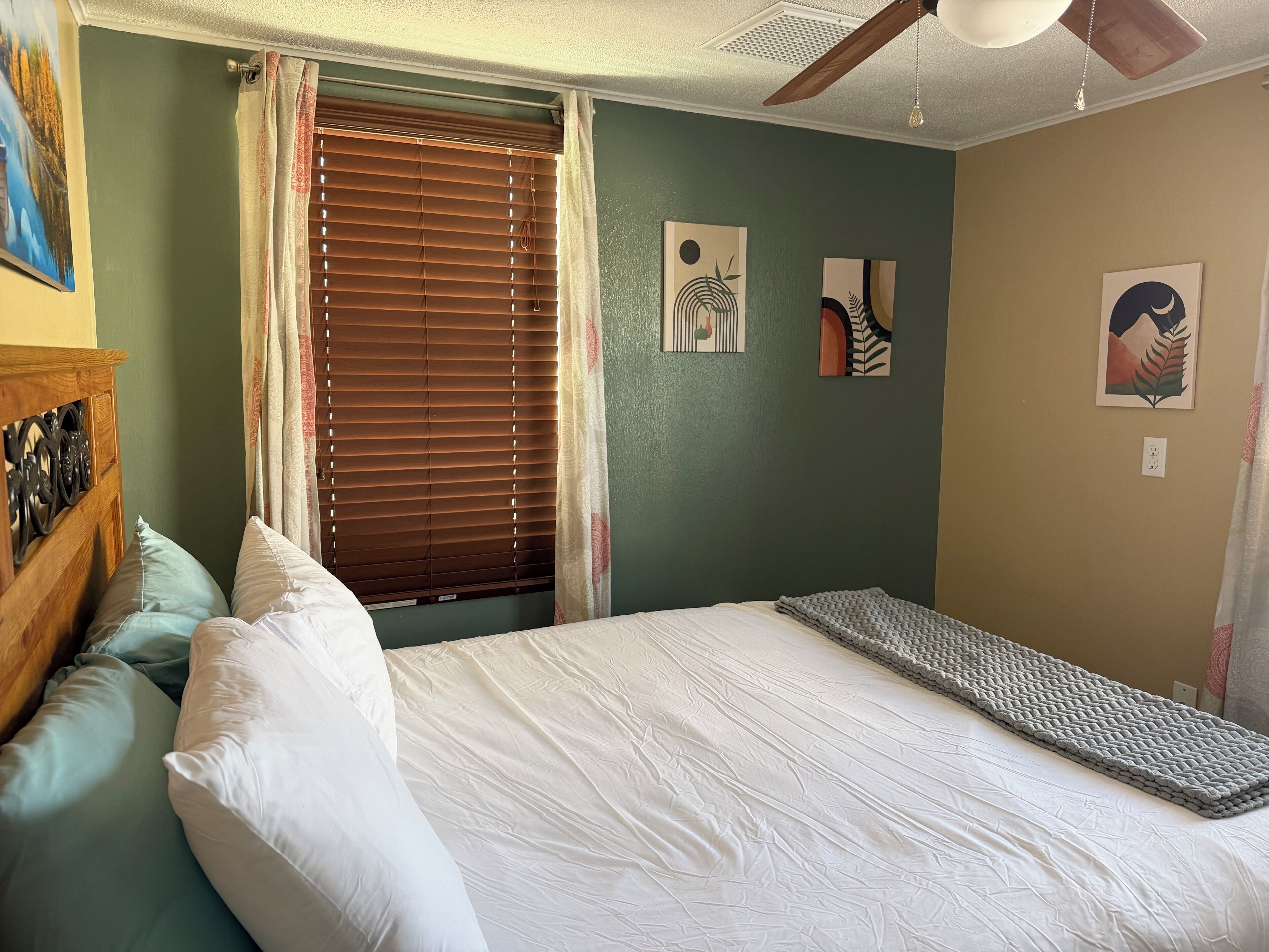 A bedroom with a bed, pillows, a headboard, a window with blinds, and wall art.