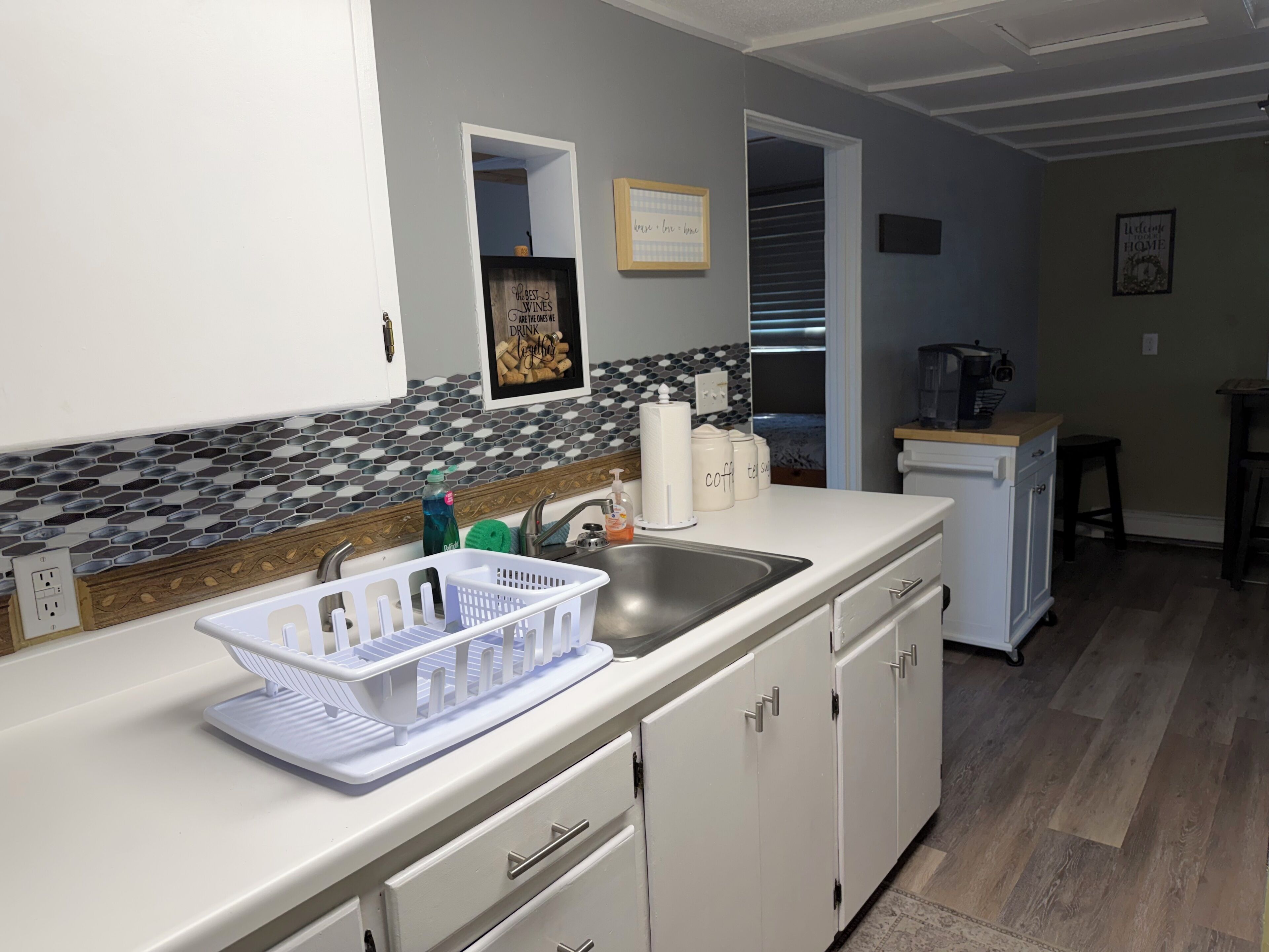 A kitchen with a white countertop, a stainless steel sink, and a tiled backsplash. There are cabinets, a coffee machine, and a refrigerator in the background.