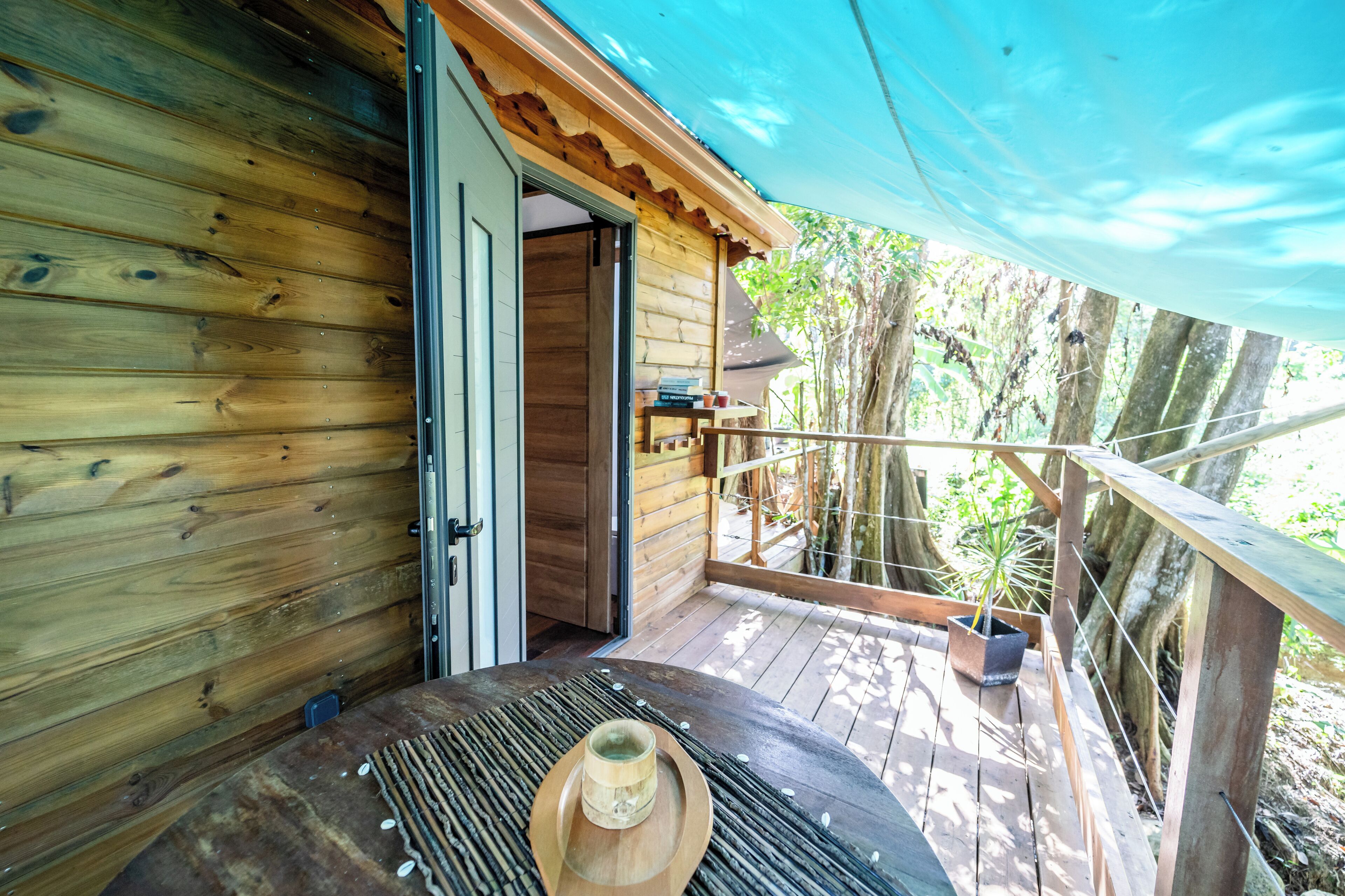 A wooden cabin with a wooden deck, a door leading inside, and a small outdoor area with a table and a hat.