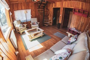 Een gezellig chaletinterieur met een houten vloer, een bank, een salontafel, twee stoelen, een lamp, een ladder en een klein rode plankje.