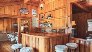A rustic bar area with wooden walls, a wooden counter, and stools with fluffy cushions.