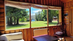 A cabin with a large window offering a view of a grassy area and trees. There is a wooden table and a chair with a cushion.