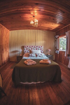 Een knus vertrek met houten wanden en plafond, een bed met bloemenbedrukking en een raam met gordijnen.