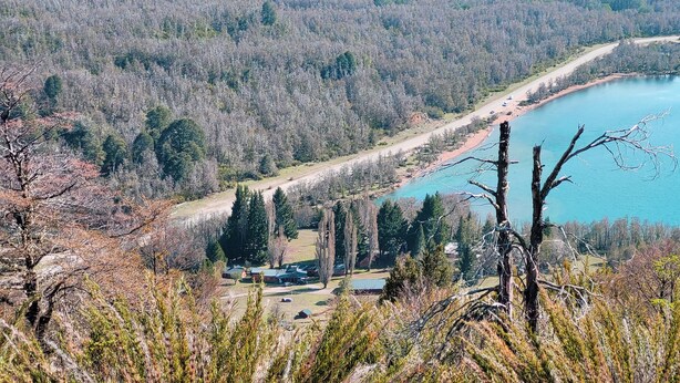 A lakeside property with a clear view of the lake, surrounded by trees and a small building with a red roof. - Hosteria Villarino (Nahuel Huapi National Park)