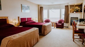 A hotel room with two beds, a desk, a chair, a fireplace, and a large window. - New Imperial Suites (Whitecourt)