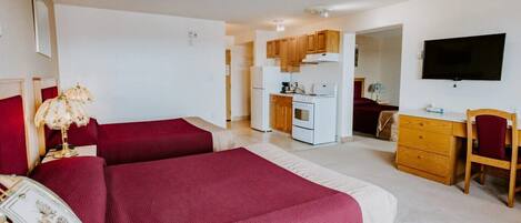A hotel room with two beds, a kitchenette, a TV, and a desk with a chair.