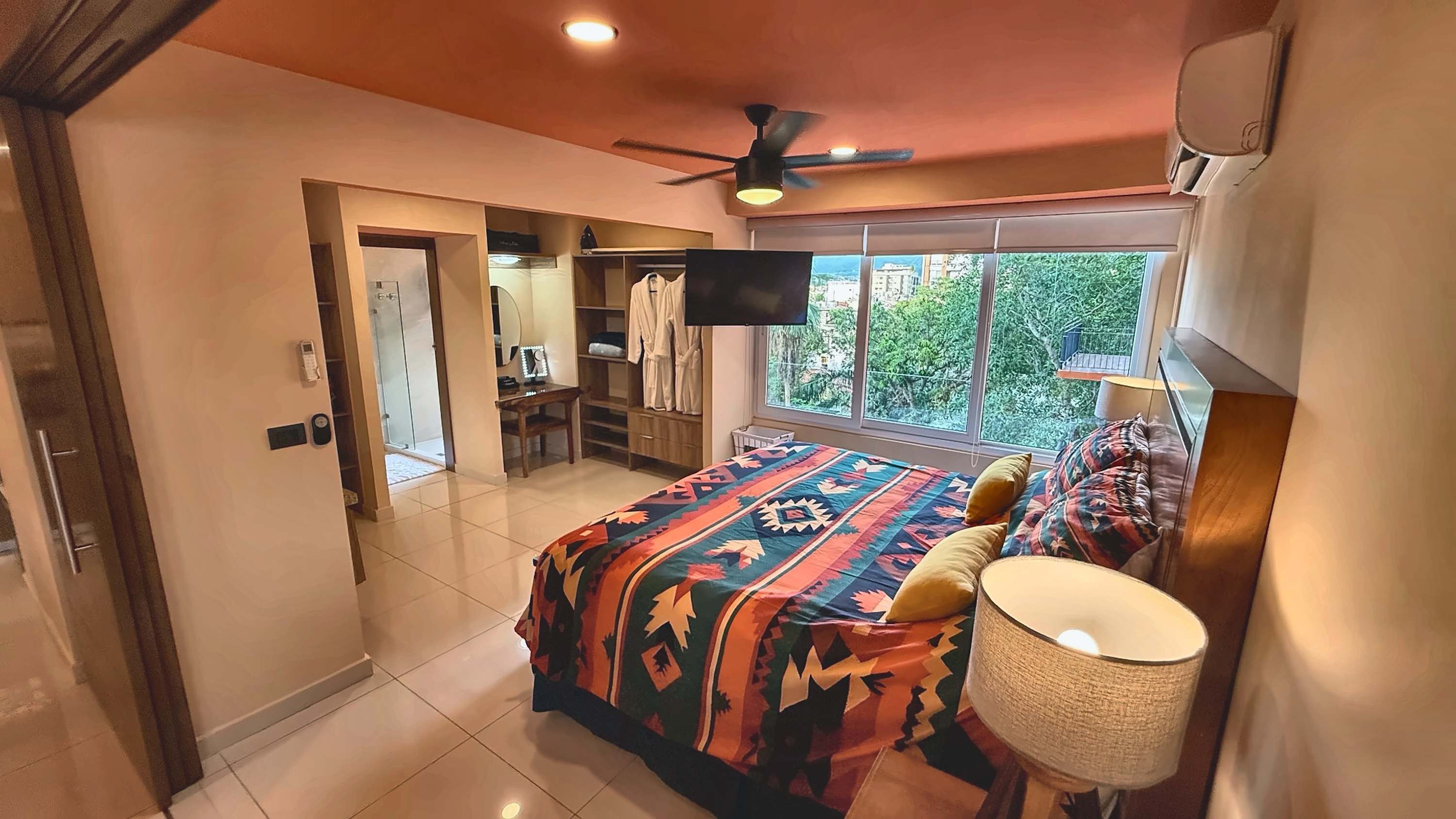 A bedroom with a bed, a TV, a ceiling fan, and a window with a view of greenery.