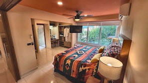 A bedroom with a bed, a TV, a ceiling fan, and a window with a view of greenery.