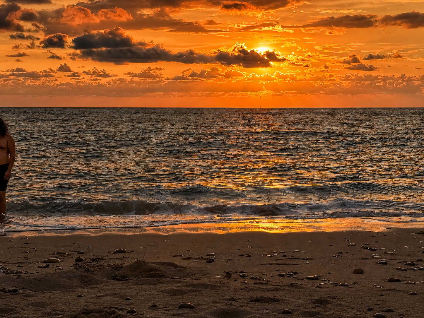 A beach with a sunset, waves, and a person standing on the shore.