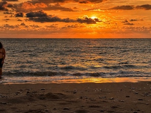 A beach with a sunset, waves, and a person standing on the shore.