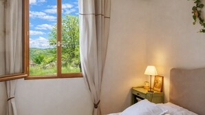 A bedroom with a bed, a nightstand with a lamp, a window with curtains, and a view of greenery outside. - Casa Di Babbo (Olmi-Cappella)