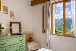 Quarto com cômoda verde, espelho, quadro emoldurado, almofada e janela com vista para as montanhas.