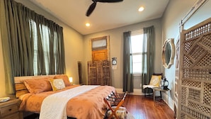 A bedroom with a wooden bed, a nightstand, a chair, a small table with a lamp, and a decorative wall panel.