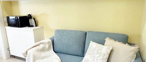 A living room with a blue sofa, two pillows, a white blanket, and a white side table with a microwave.