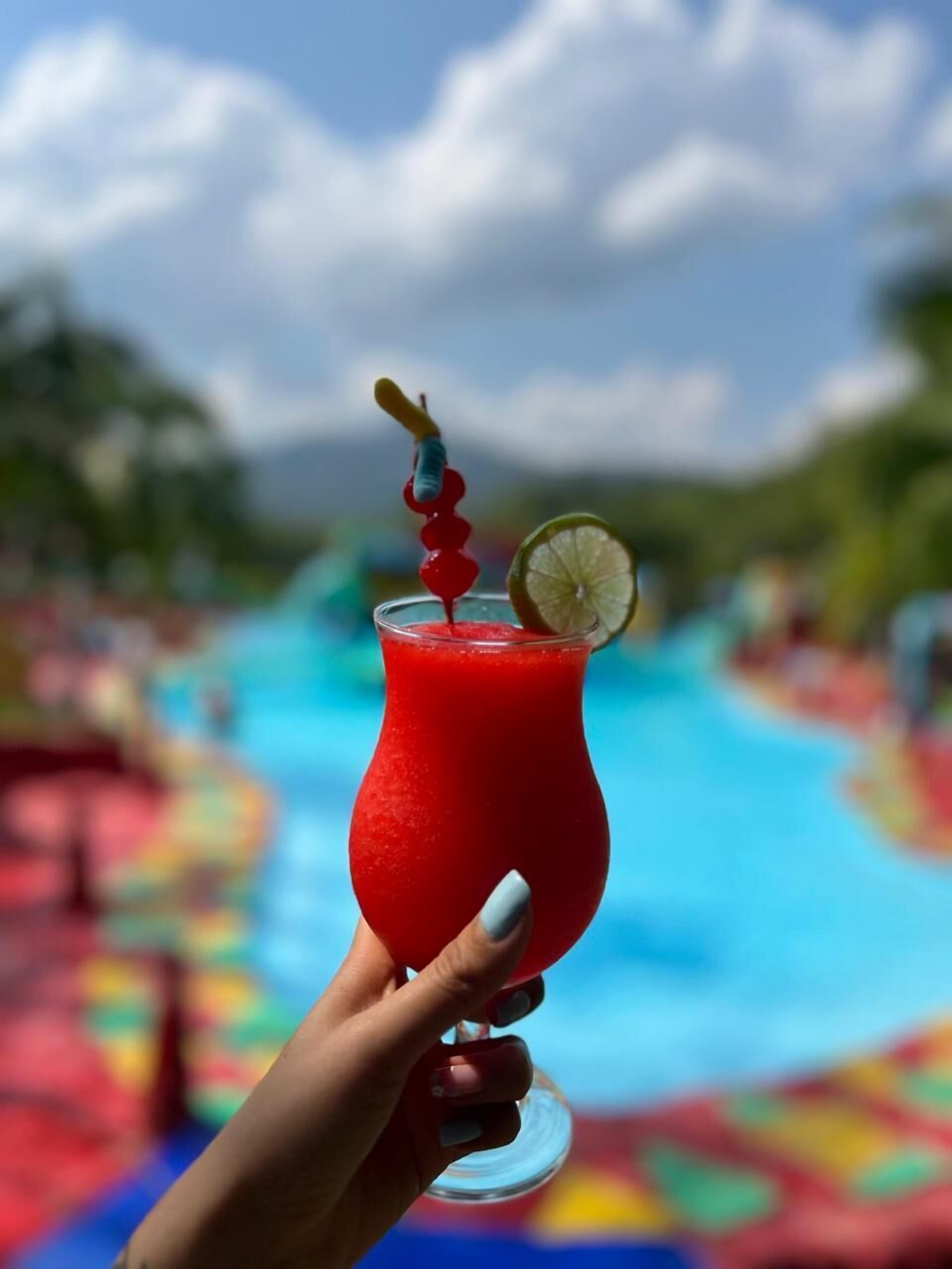 Una mano sosteniendo un vaso con una bebida roja, una cuña de lima y un adorno, frente a una piscina con un deck colorido.